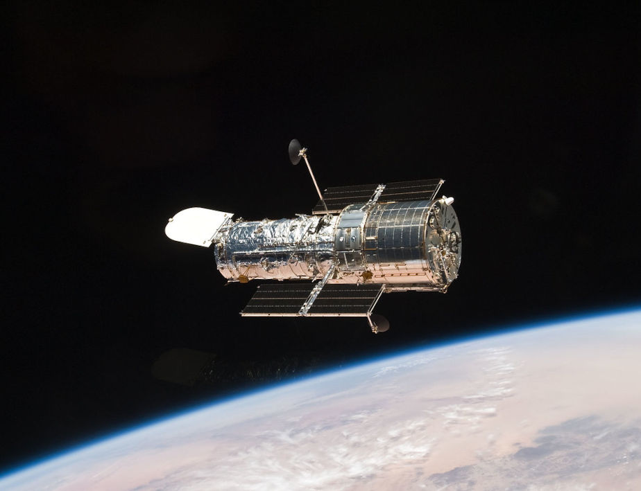 The colored image shows the silvery Hubble telescope floating in space in the center. The Earth and Earth's atmosphere can be seen in light blue below.