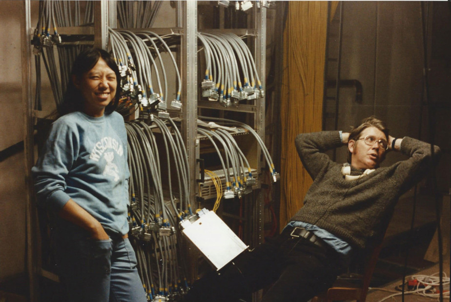 The photo shows two people: on the left stands Sau Lan Wu wearing a light blue sweatshirt. On the right sits Bjørn Wiik in a brown knitted sweater with his arms folded behind his head. In the background you can see many gray cables hanging out of the wall and in the middle of the picture a clipboard with a white sheet of paper on it.
