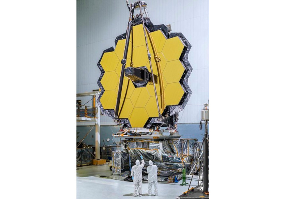 The colored image shows the mirror of the James Webb telescope in a large hangar. Two people in white protective suits stand in front of it.