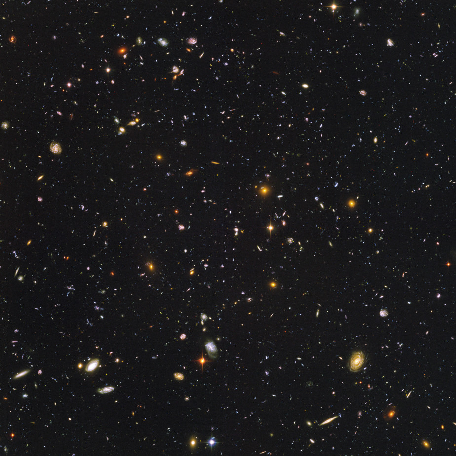 Die farbige Abbildung zeigt die Weiten unseres Universums. Es sind sehr viele kleine Punkte zu sehen. Ein paar Punkte sind größer und leuchten in orange, blau, lila, rot und gelb. Bei ein paar der größeren Punkte kann man die spiralförmige Struktur einer Galaxie erkennen.