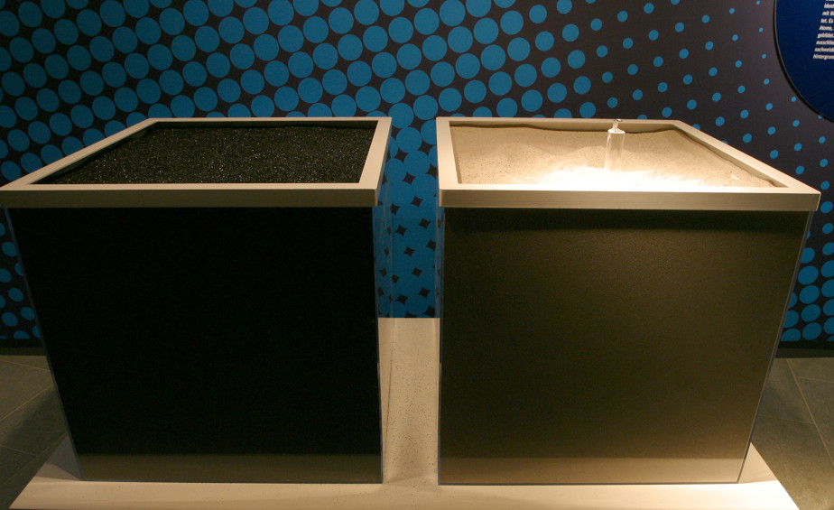 Photo of two square containers filled with sand. The sand inside the left box is black while the one on the right is light beige. On a small cylinder sticking out of the light sand, a single grain of sand is laid out separately.