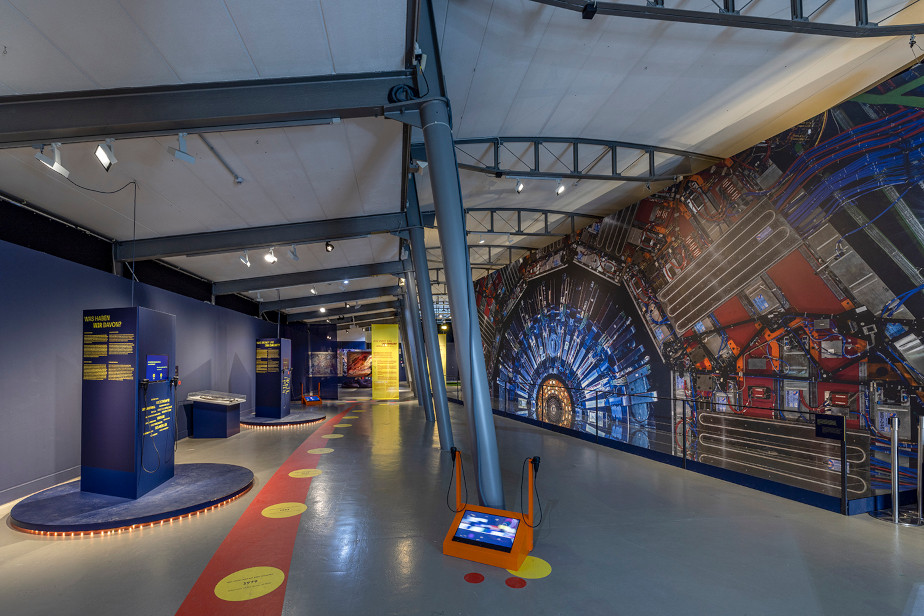 Photo from the exhibition: the right half of the image is occupied by a large screen showing the cross-section of the CMS detector in blue, silver and red. The rest of the photo shows other exhibits on the left and gray columns supporting the ceiling in the center. The image of the CMS detector looks huge in comparison.