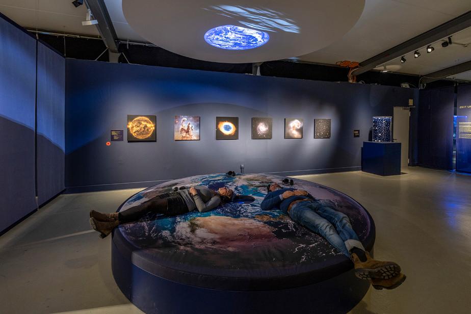 Das Foto zeigt im Vordergrund eine kreisrunde Liegelandschaft, auf der zwei Menschen mit Kopfhörern liegen und nach oben schauen. Über ihnen an der Decke hängt eine ebenfalls runde Leinwand, die gerade ein Bild von der Erde über dem Mondhorizont zeigt. Im Hintergrund hängen an einer blauen Wand Bilder des Hubble Weltraumteleskops.
